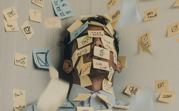 The image represents someone feeling stressed. Someone tilts their head back into a corner and they have post-it notes stuck on them and on the walls.